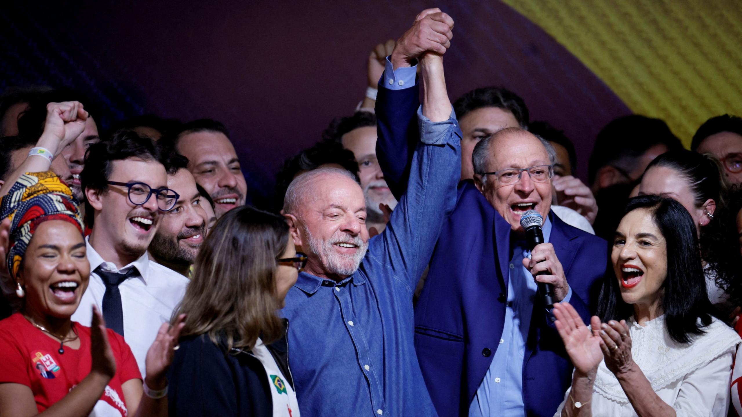 Lula smiles as his vice-president clasps and holds his hand into the air, speaking into a microphone. A crowd of people are cheering, clapping and smiling around them.